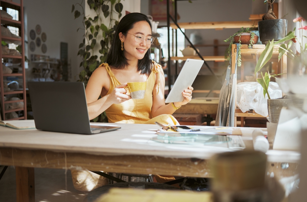 Woman in yellow dress holding a tablet and coffee cup, sitting at a desk with a laptop, surrounded by art supplies in a creative workspace, illustrating brand development for small businesses.